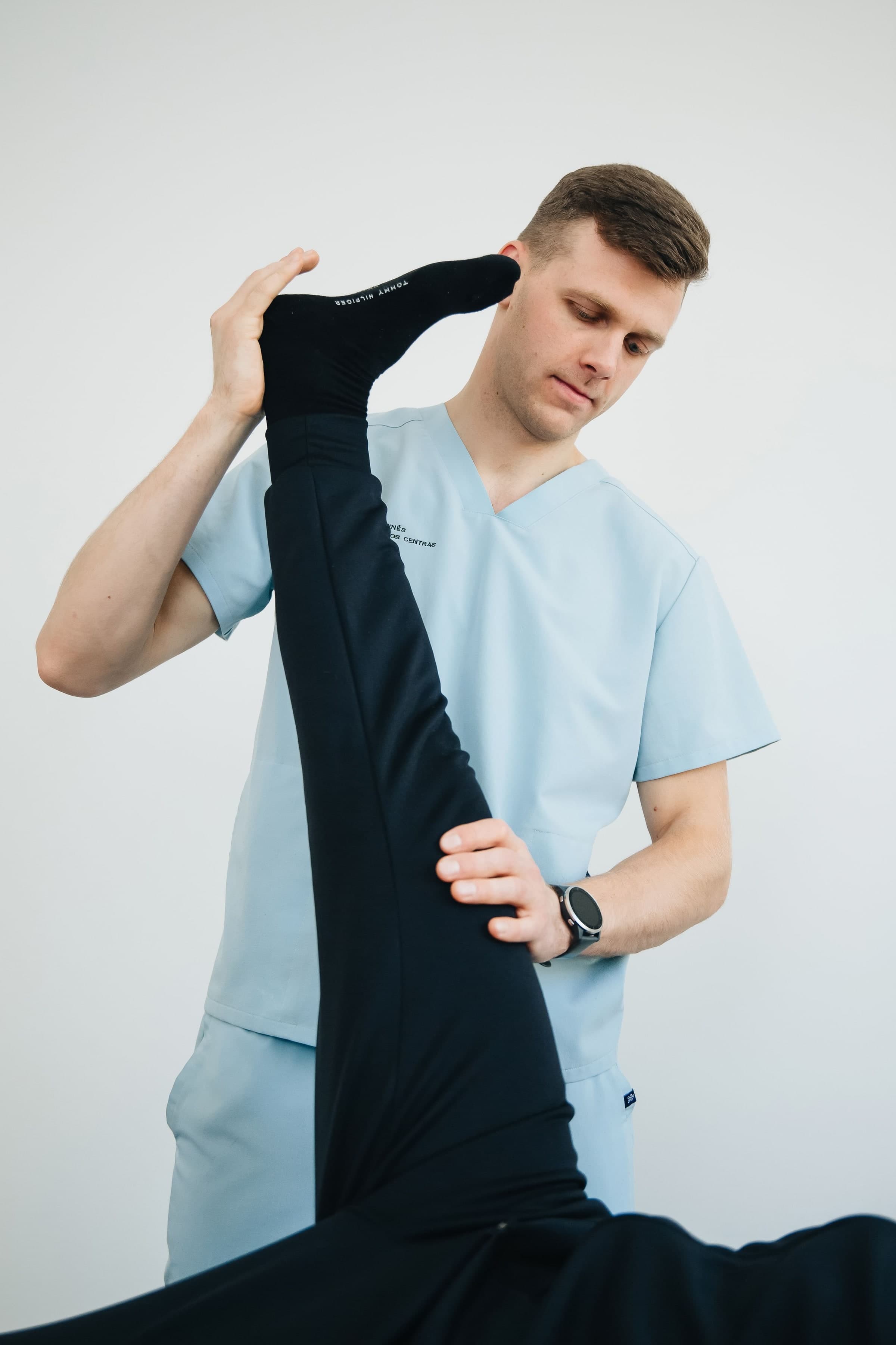 A healthcare professional in a light blue v-neck uniform demonstrating a stretching technique by holding a black compression sleeve along their leg, Toronto