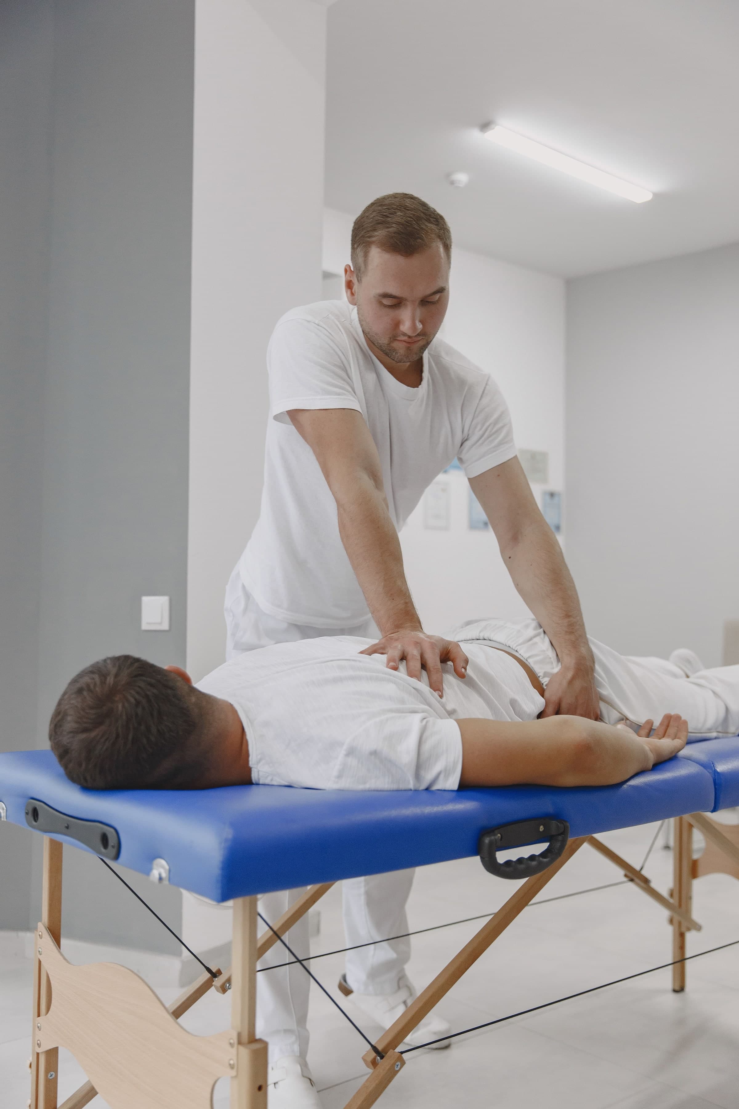 A male massage therapist performing a back treatment on a patient lying face-down on a blue portable massage table in a bright white clinical room in Toronto